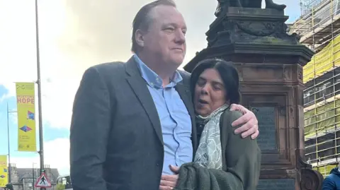 A man in a grey suit jacket and a open-necked blue shirt hubs a smaller woman, who is wearing a green jacket and a green, white and black scarf. She has her eyes closed and he is looking off to the left of the camera. The base of a statue, lamp posts and scaffolding around abuilding can be seen in the background.