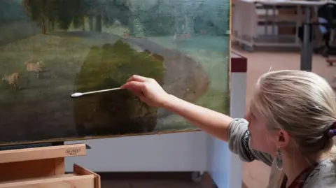 National Trust Image of a woman with a blonde ponytail, who is holding a long cotton bud and using it to clean and rsetore the corner of a large oil painting on an easel.