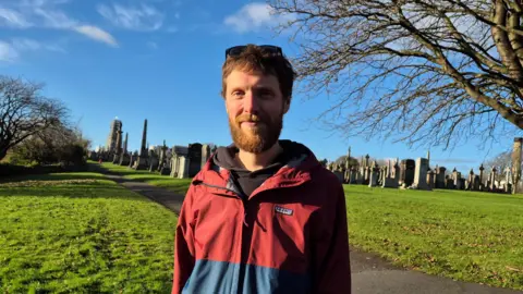Greg Hawke, who has red hair and a beard, standing outside the graveyard on a sunny day. He is wearing a red and blue jacket, with sunglasses on top of his head. 