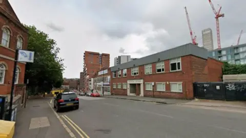 A city street with a mix of older and modern buildings. On the left, there is a red-brick building with arched windows and a sign. In the center, a two-story brick building labeled “Kendal House” has a flat roof and rectangular windows. A black car is parked on the left side of the road. In the background, there are high-rise buildings and several red construction cranes.