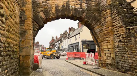 A roman bricked archway. The sandy coloured bricks are chipped in places. Green moss covers patches of the monument. The street can be seen through the archway, red bollards cover sides of the streets. A tractor can be seen in the background lifting supplies.