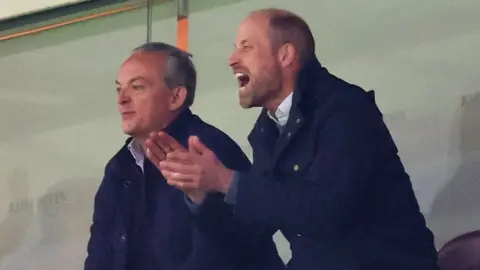 Getty Images Prince William pictured with his clapping his hands and shouting, with another man stood next to him slightly smiling.