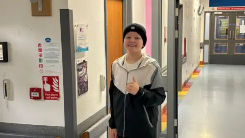 Chloe Louie pictured with a beanie hat on and his thumb up on a hospital corridor.