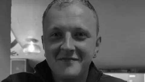 A black and white image of Daniel Sowerby. He has short hair and an earring on his left ear. He is smiling at the camera.