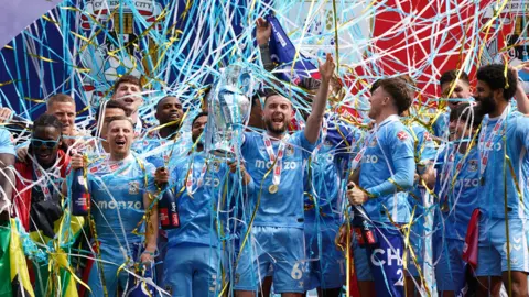 The Championship-winning Coventry City side are covered in streamers after lifting the trophy. Club captain Matt Grimes is in the mddle of the picture with the Championship trophy in his right hand and a medal around his neck.
