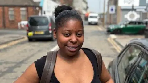 A young, black woman with hair tied in a bun. She is wearing a backpack. 