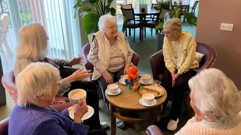 Monica Wingrove sits in a circle surrounded by friends drinking coffee and easting biscuits. On the table is a red collecting tin in the shape of a jelly baby. 