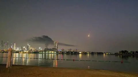 Reuters A shot of Dubai's skyline with smoke rising from a high-rise building - the hotel Carson was staying in. It is nighttime and a body of water is reflecting the city lights. The streak of a missile can be seen in the distance.