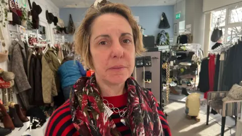 Emma Kemsley standing in her shop surrounded by clothes, shoes and jewellery stands. She is wearing a black and red striped jumper, a red patterned scarf and a spherical silver necklace. She has short dirty blonde hair and a forlorn expression. 