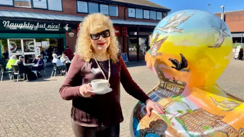 A woman with long, blonde, curly hair, a maroon long-sleeved top and black leather trousers is holding a white cup of coffee and saucer. She stands in a town square beside a giant painted duck. The duck is decorated with images of birds, historic buildings and a sunset. There is a cafe/restaurant behind her with people sat outside drinking. There are office units above and two more adjoining shops.