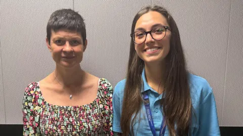 Two NHS Somerset nursing apprentices are standing next to each other in front of a white wall. The left woman is wearing a colourful dress and has short hair. The woman on the right is wearing a blue shirt and has long, dark hair.