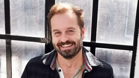 Head and shoulders image of a smiling Alfie Boe standing in front of a window with small black framed panes. He has short dark hair and a beard and is wearing beige button up top under a navy shirt jacket