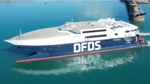 BBC A picture of a large blue body of water with part of the harbour visible. There is a large white and blue boat with DFDS written on its side.