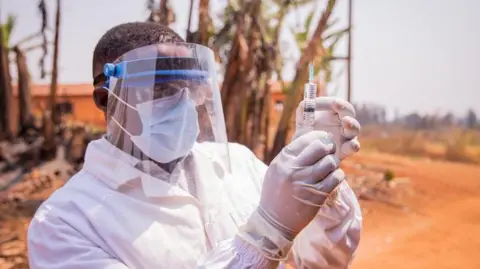 A medic in full protective face mask and white clothing, standing outside, holding up a syringe with gloved hands 