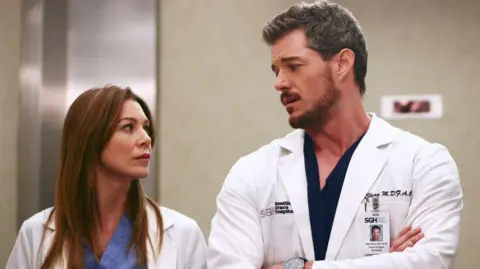 Eric Dane and Ellen Pompeo in a still from Grey's Anatomy. Both wear white medical coats over light blue and black scrubs.
