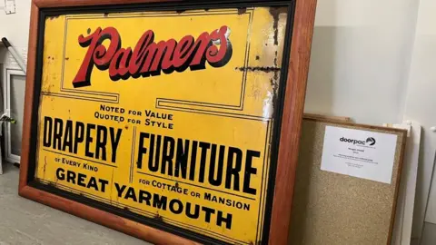 Andrew Turner/BBC Yellow enamel sign, in a wooden frame, with black text, stating:
PALMERS
NOTED FOR VALUE; QUOTED FOR STYLE
DRAPERY OF EVERY KIND
FURNITURE FOR COTTAGE OR MANSION
GREAT YARMOUTH
