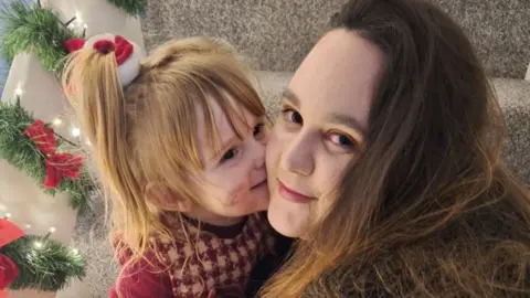 Family photo A woman with reddish brown long hair being kissed on the cheek by her toddler daughter. The little girl is blonde with a pony tail and is wearing a red and white dress over a red sweater. The mother and daughter are pictured on the stairs and and there are Christmas decorations on the handrails