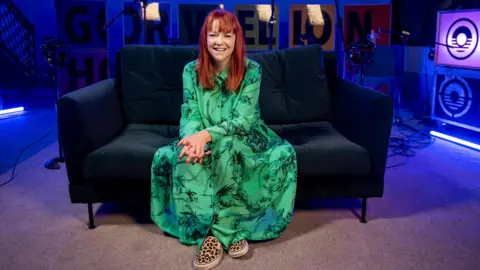 Radio Wales presenter Bethan Elfyn is sat on a sofa and smiles at the camera. the background is a studio set. She is wearing a green dress and has red hair