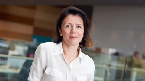 Prof Samra Turajlić, director of the Cancer Research UK Manchester Institute, with shoulder length brown hair wearing a white short-sleeved shirt and gold earrings. She is standing in a research lab. She is smiling.