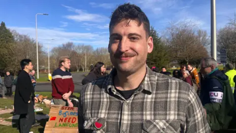Matt Riemland smiling straight at the camera. He has dark hair and a short dark moustache and dark stubble. He is wearing a checked shirt and is standing in front of a crowd of people.