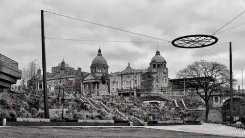 Stewart Paul A circle structure hangs from ropes above a park. An ornate building with domes is in the background. It is a black and white picture.