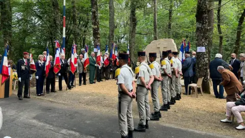 Joanna Burri-Weaver Representatives of the French army and veterans' organisations stand in two rows by a large stone monument in a forest
