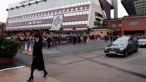 PA Media A funeral procession, led by a woman wearing a black hat with a veil down the back, makes its way past Villa Park with crowds gathered behind a barrier outside the stadium. The woman is followed by two black cars.
