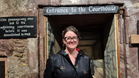 A woman standing inside an old building. She has dark hair and glasses, and is standing in front of a sign saying 'entrance to the courthouse'