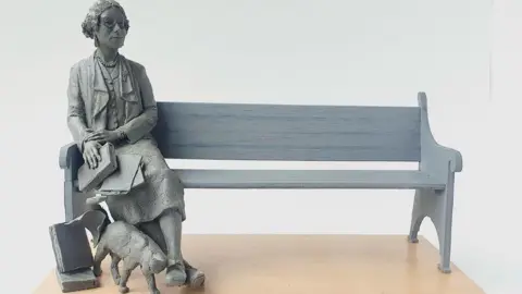 Denise Dutton/Dorchester Civic Society A clay maquette of the statue depicting a woman sitting at one end of a bench with a cat rubbing its head on her feet. The woman is wearing a long skirt and a buttoned blouse. She is also wearing glasses and has curly hair parted in the centre.