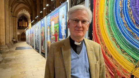 The Rev Canon Jonathan Baker - a man with grey hair and round spectacles - is wearing a clerical collar, a blue jumper and tweed blazer. A minster's passage can be seen behind him along with a colourful display alongside the passage depicting different biblical scenes.