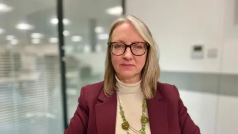 Martin Giles/BBC Caroline Topping is sitting in an office with blinds in the background and is wearing a maroon suit jacket and camel coloured roll neck jumper underneath. She has a jade green beaded necklace and glasses. 
