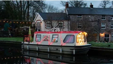 Steve Hale Photograph of a narrow boat along Brecon and Monmouthshire canal. A 225 sign sits on top of the narrow boat, and red fairy lights zig zag up its side. In the background, a stone wall cottage can be seen set up from the grass behind the canal, and a row of fairy lights are attached to the garden wall on the left. 