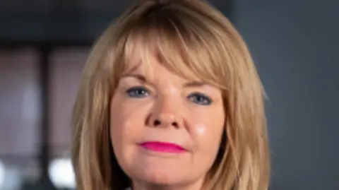 Monica Fogarty faces the camera. She wears pink lipstick and has a blonde bob haircut.