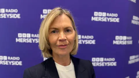BBC News/Claire Hamilton A blonde woman looks directly at the camera. She has bobbed hair, dark blue eyes and a slight smile. She's wearing a dark jacket and white t-shirt and is standing in front of a BBC Radio Merseyside banner. 