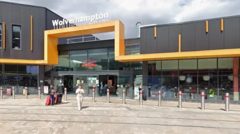 Google A metal, yellow bar runs along the front of the station a few metres off the ground and goes higher and forms a square shape above the glass door entrance to the station. Bollards are across the entrance and glass windows either side as some passengers are coming out.
