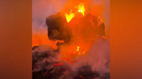Shropshire Fire and Rescue Service A large farm vehicle carrying bales of hay that is completely engulfed in orange flame and red hot fire. There are flames and smoke billowing from it