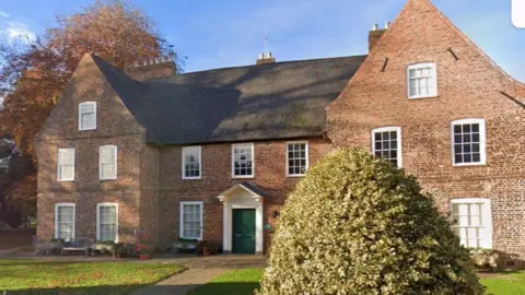 A large, brick-built manor house with thatched roof sits in a landscaped garden, with trees, bushes and lawns.