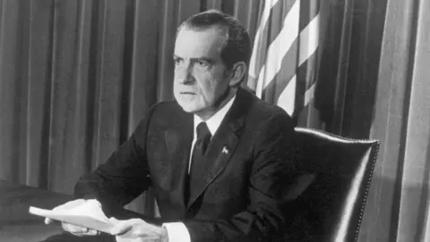Getty Images Black and white image of Former US President Richard Nixon taken during live broadcast of resignation in 1974. He is wearing a suit, clutching papers and sitting at a desk.