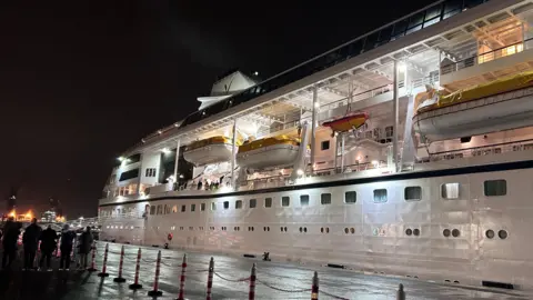 The ship has three stories and lots of small windows.  The sky is black and there are lights on in the windows.   Passengers are visible on the ship. 