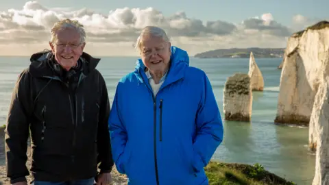 Silverback Films Keith Scholey left is wearing a black mac and smiling at the camera. He has grey hair and is wearing glasses. Next to him is Sir David Attenborough, also smiling and with his hands in the pockets of a bright blue raincoat. They are standing in on coastal headland with the sea behind them.