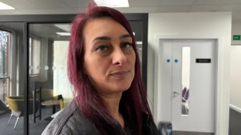 Martin Heath/BBC Josephine Quinton with long dark red hair wearing a black leather jacket and looking at the camera in an office building with a white door to her left and chairs and tables to her right.