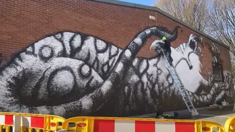 Sarah Jay Hawley Phlegm, who is wearing a lime green beanie hat, is at the top of a ladder creating the artwork - here he's working on the last portion, the cat's face.