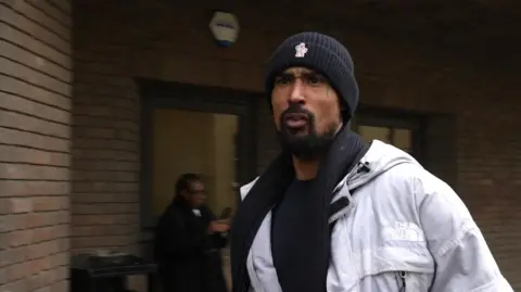 Ashley Warren walks outside the bricked frontage of Chelmsford Crown Court, wearing a white coat and dark-coloured beanie hat. He is mouthing something towards people behind the camera.
