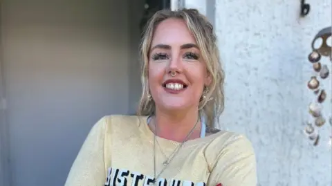 A woman in a yellow jumper with 'sisterhood' written on the chest is standing in front of her home and smiling into the camera. She has blonde hair tied in a ponytail and is wearing jewellery including a long silver necklace, gold earrings and has a nose ring.