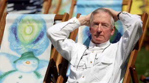 PA Media Author Raymond Briggs sat on a deck chair. He the deck chairs have drawings of The Snowman on them.
