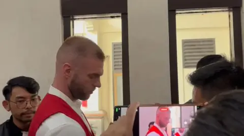 Thomas Parker leaving court. He is wearing the red waistcoat given to defendants. He is surrounded by a number of people. A man in the foreground of the picture is filming Parker on a mobile phone.