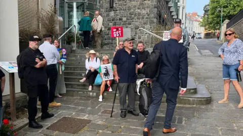 Eleven protestors outside the States with three people holding no to GST placards. A man, Deputy David Dorrity who we see from behind is walking up the stairs to the Royal Court. 