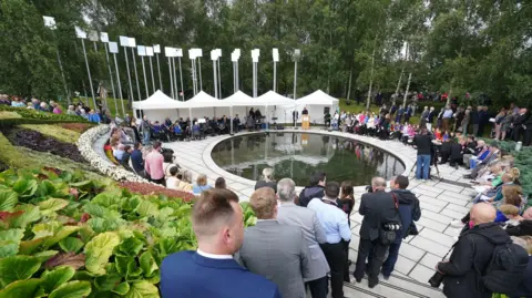 Brian Lawless/PA Wire A view of the Memorial Gardens in Omagh during a 2023 ceremony to mark the 25th anniversary of the bombing.  People are sitting and standing around a circular pond, flanked by 31 poles topped with mirrors to reflect light - the poles represent the 29 people who died as well as a set of unborn twins.