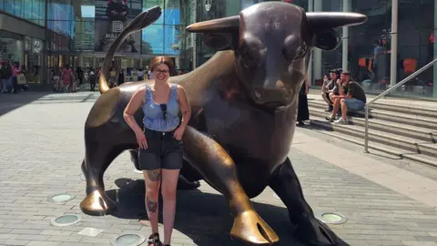 Beck Evans A person stands beside a large bronze bull statue outside a modern shopping complex with glass walls and a curved canopy roof. The person is wearing a sleeveless patterned top, dark shorts, and sandals, and has visible tattoos on one leg. Behind them, the building façade features a large Nike swoosh logo, and people are sitting on steps or walking through the open plaza. Bright sunlight casts strong shadows across the paved ground.
