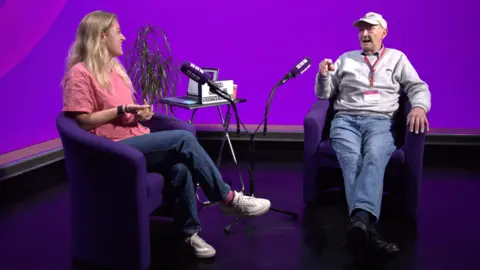 Anya Culling and Malcolm Metcalf are sitting in purple chairs in a TV studio with a purple pattern on the screens behind them, which is reflecting off the black floor. Culling has long blonde hair and is wearing a pink t-shirt, with blue jeans and white trainers. Metcalf is sat to her left, with two microphones on stands and a plant and table between them. He is wearing a white baseball cap, a grey jumper, blue jeans and black shoes.